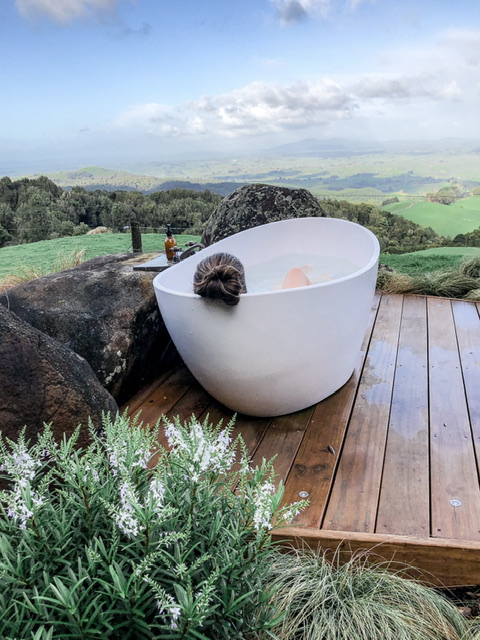 A young woman relaxing in a large outdoor white bath photographed from behind with only brunette hair in a bun and her right knee visible. Views of a green farm below are visible.
