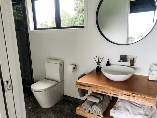 Bathroom inside Rua Hiwi Hut in the Waikato with white toilet and basin set in wooden vanity. Bathrobes and rolled towels are stashed under sink.