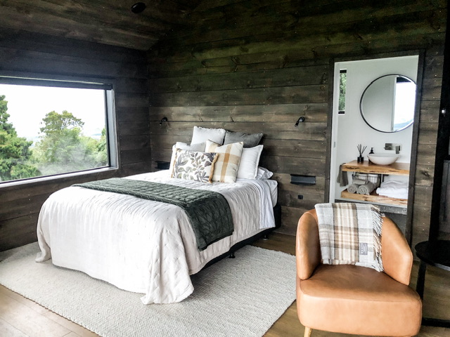 Looking into Rua Hiwi Hut through glass sliding doors. Bathroom vanity is visible through open sliding door with a brown leather armchair in the foreground and bed is on the left of the room.