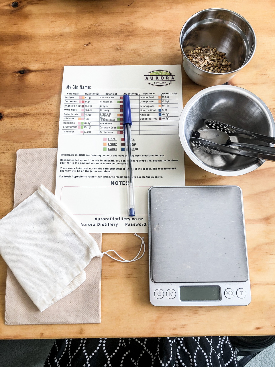 Aurora Distillery recipe card, blue pen, botanicals, scales and tools for measing gin ingredients flat laid on a wooden table.