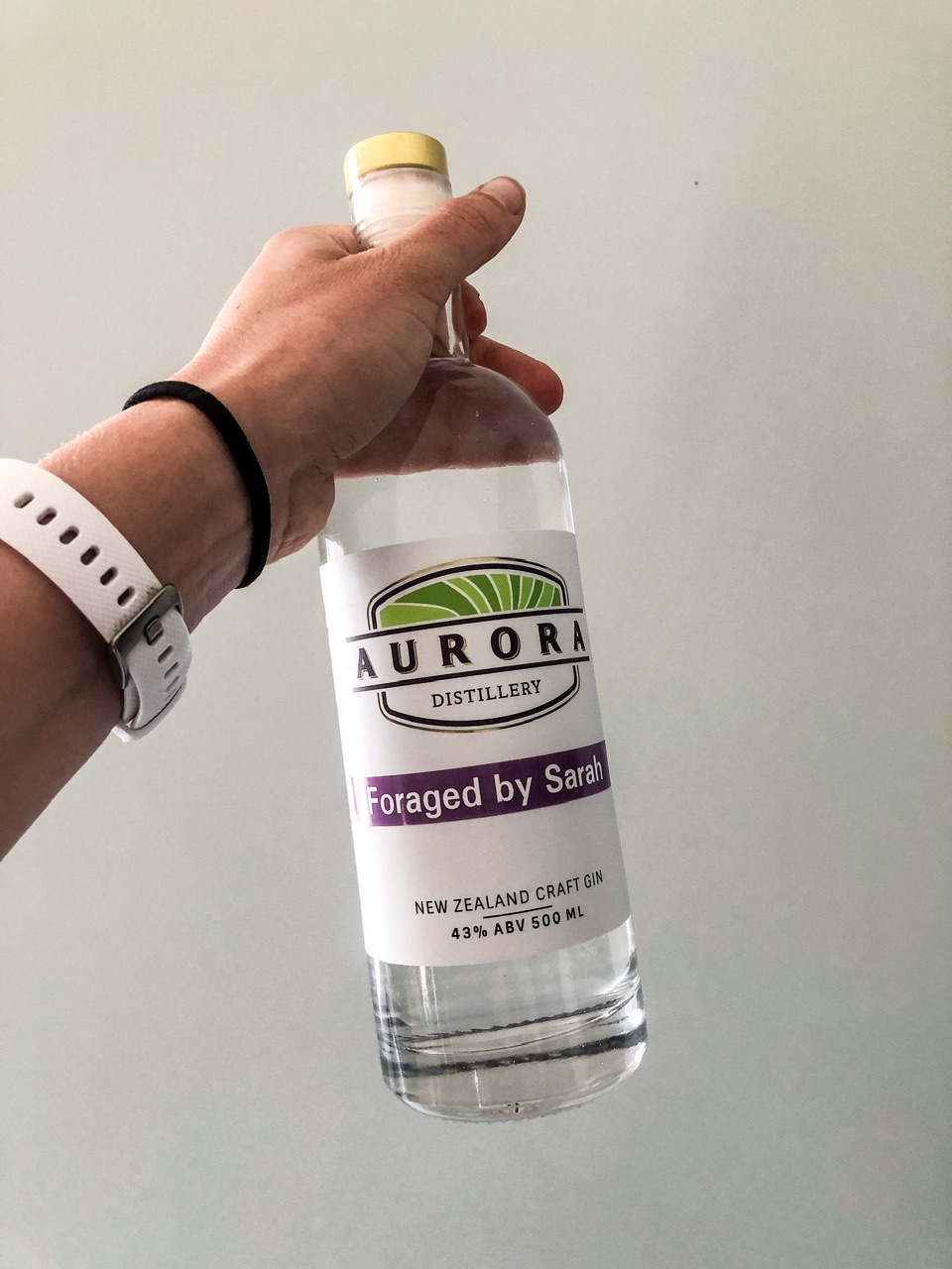 Hand holding up a bottle of Aurora Distillery Gin with the label "Foraged by Sarah". The person's wrist has a white watch strap and black hair tie around it.