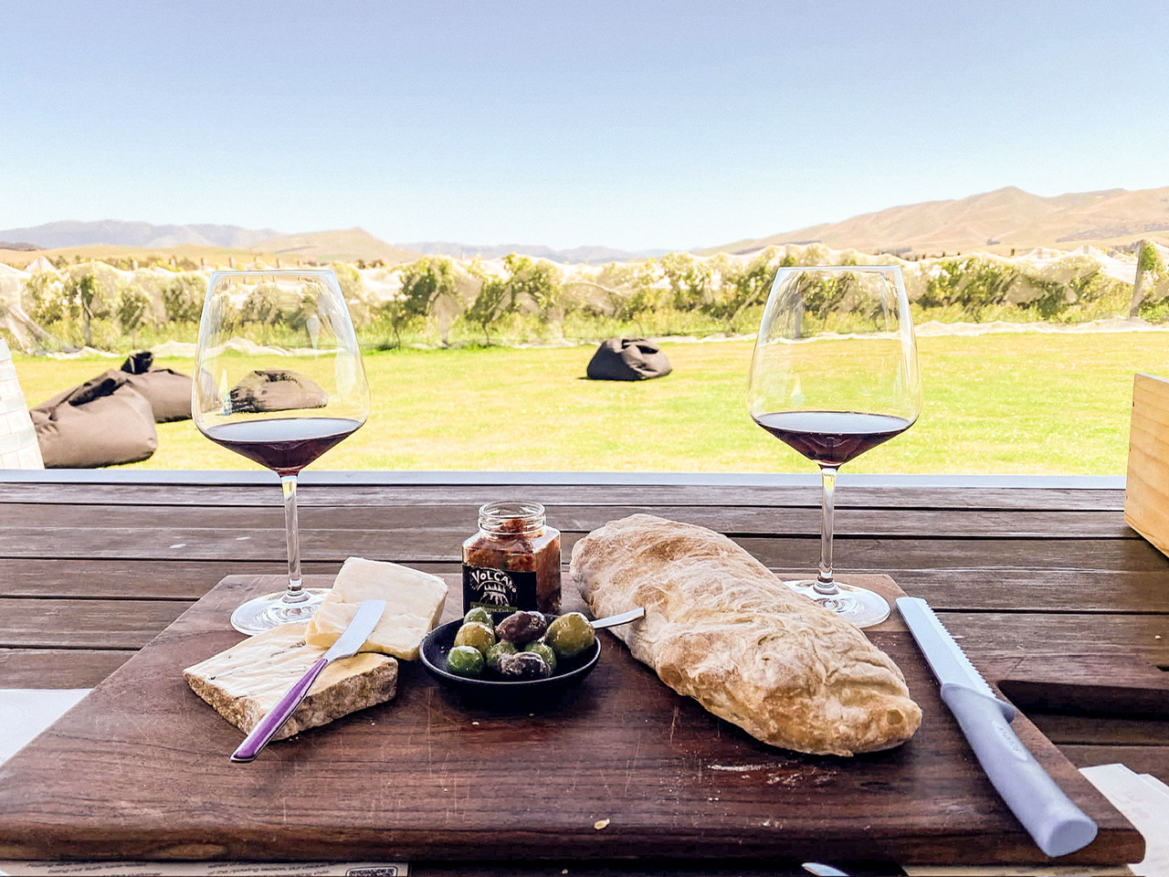 Wine tasting set up at Georges Road Wines in Waipara with a cheese platter and two glasses of red wine on a brown picnic table with beanbags on a lawn in the background with wine vines behind on a sunny day.