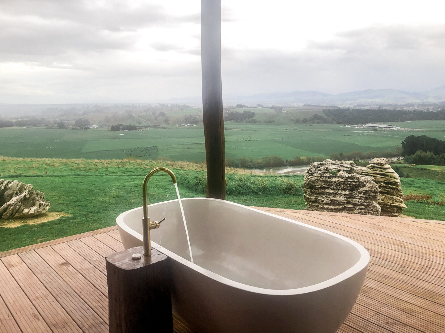 Outdoor bath at Fossil Hill Canopy Camping. A large white outdoor bath with a running tap sits on a wooden deck with views of fossils and green farmland.