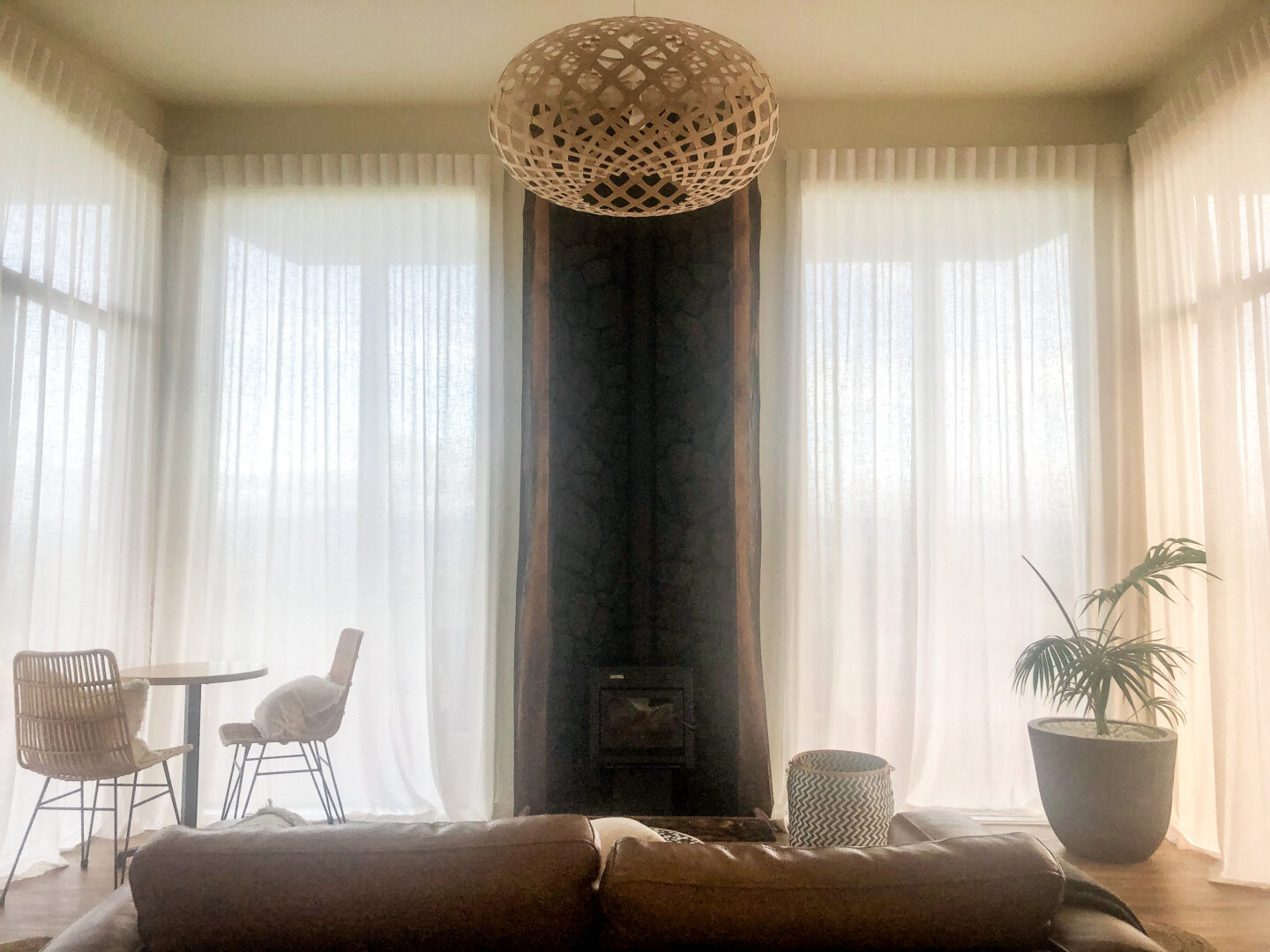 Interior of Fossil Hill Hut, a Canopy Camping site in the Wairarapa. A stone background around a fireplace sits between windows with sheer drapes . A brown couch faces the fireplace and there is a green potted fern on the right and a table with two chairs in the left corner.