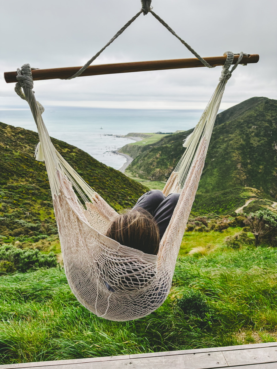 A young brunette woman lies in a hammock overlooking Cook Strait at The Ocean House Wellington.