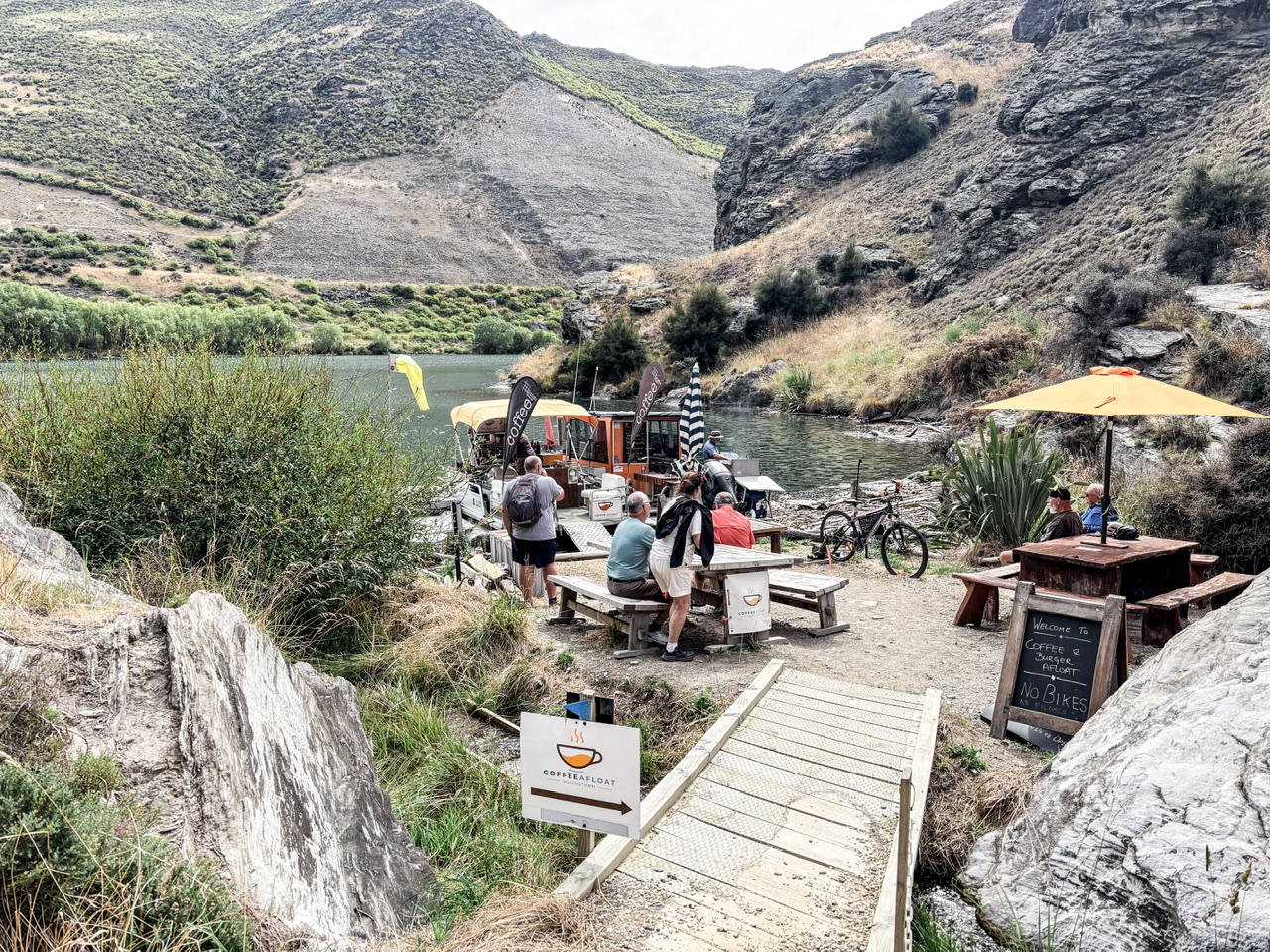 Two barges forming Coffee and Burger Afloat on the edge of Lake Dunstan. Two outdoor picnic tables are set up around 8 people are at the cafe.