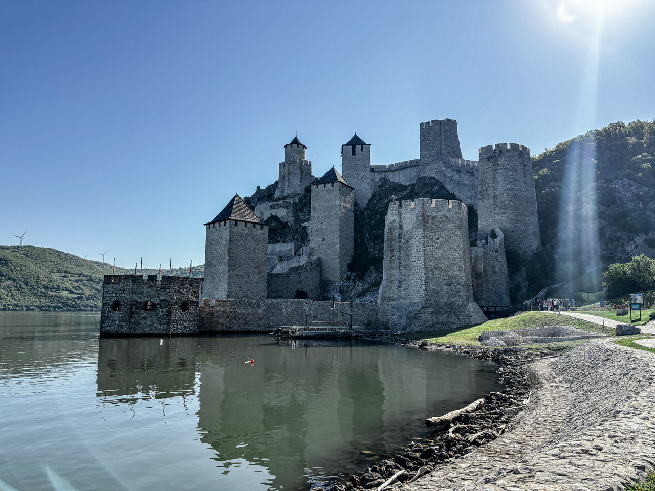 Exterior shot of Golubec Fortress in Serbia. The Danube River runs to the left of the fortress and wind turbines are visible on the on green hills on the other side of the river.
