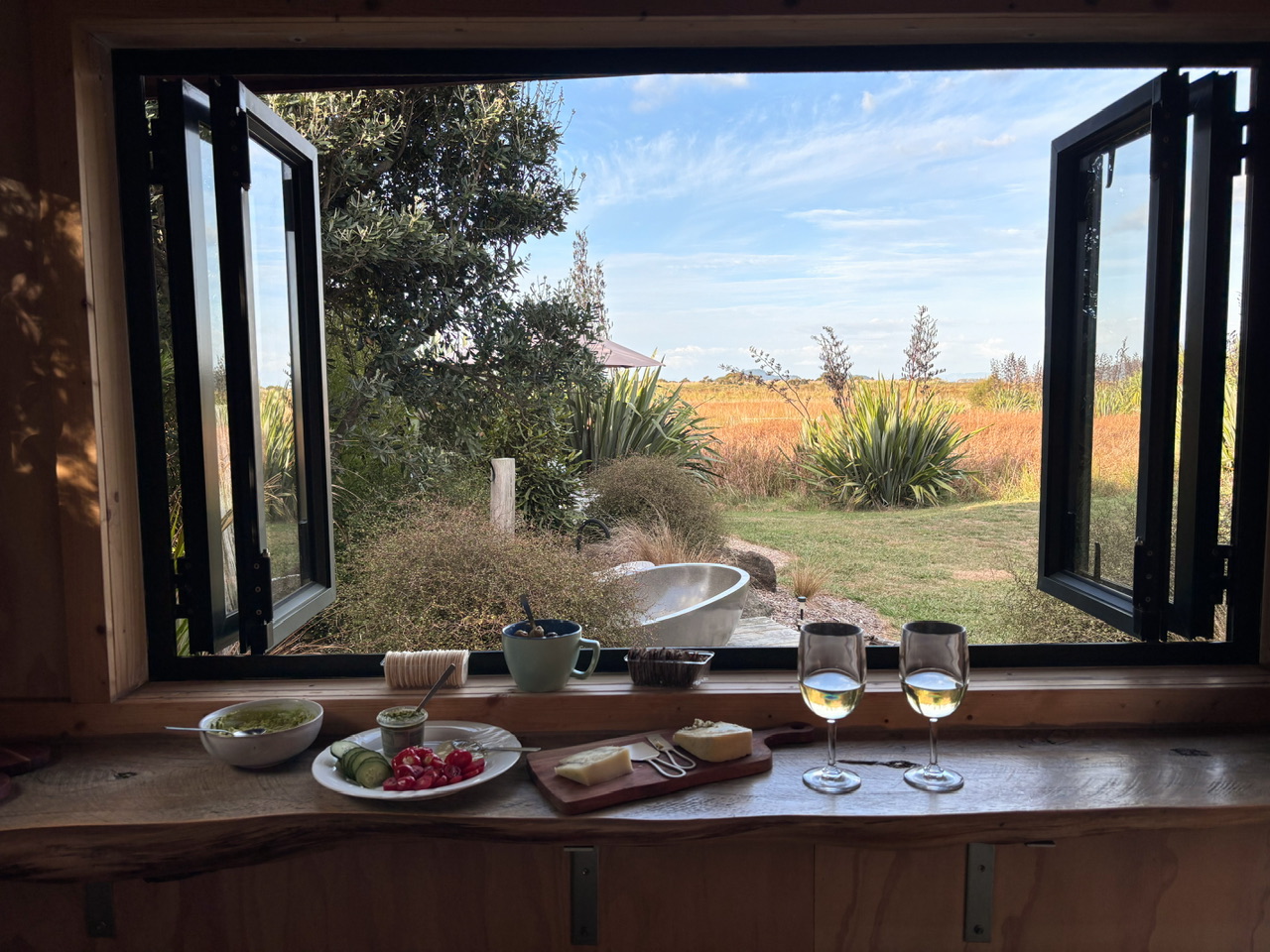 Looking out over the outdoor area including outdoor bath at Wahapū Cabin in Cleaveland. On the bench in the kitchen in front of open windows is a prepared cheese platter.